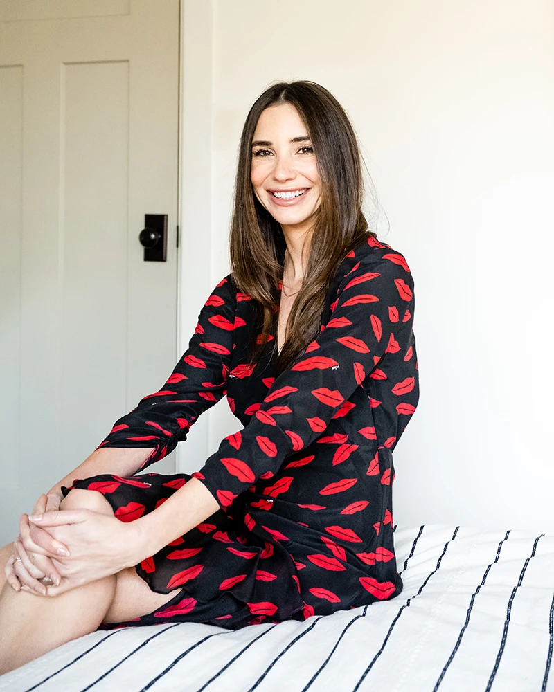 Blaine wearing a black dress with a red pattern, seated on a bed, smiling