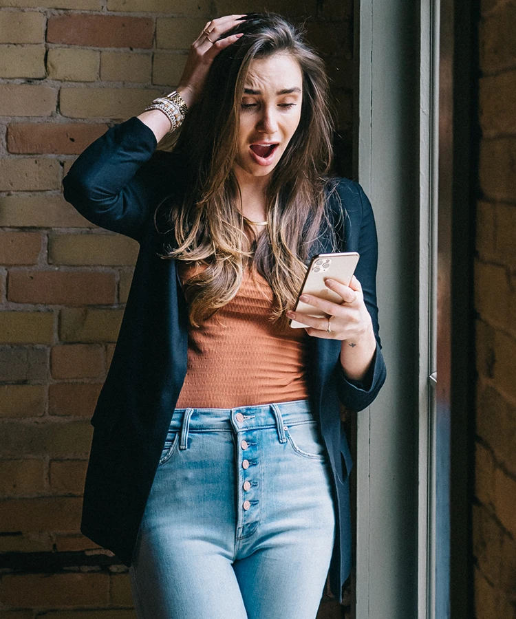 Blaine wearing a navy blazer in a brick loft holding her phone, looking surprised