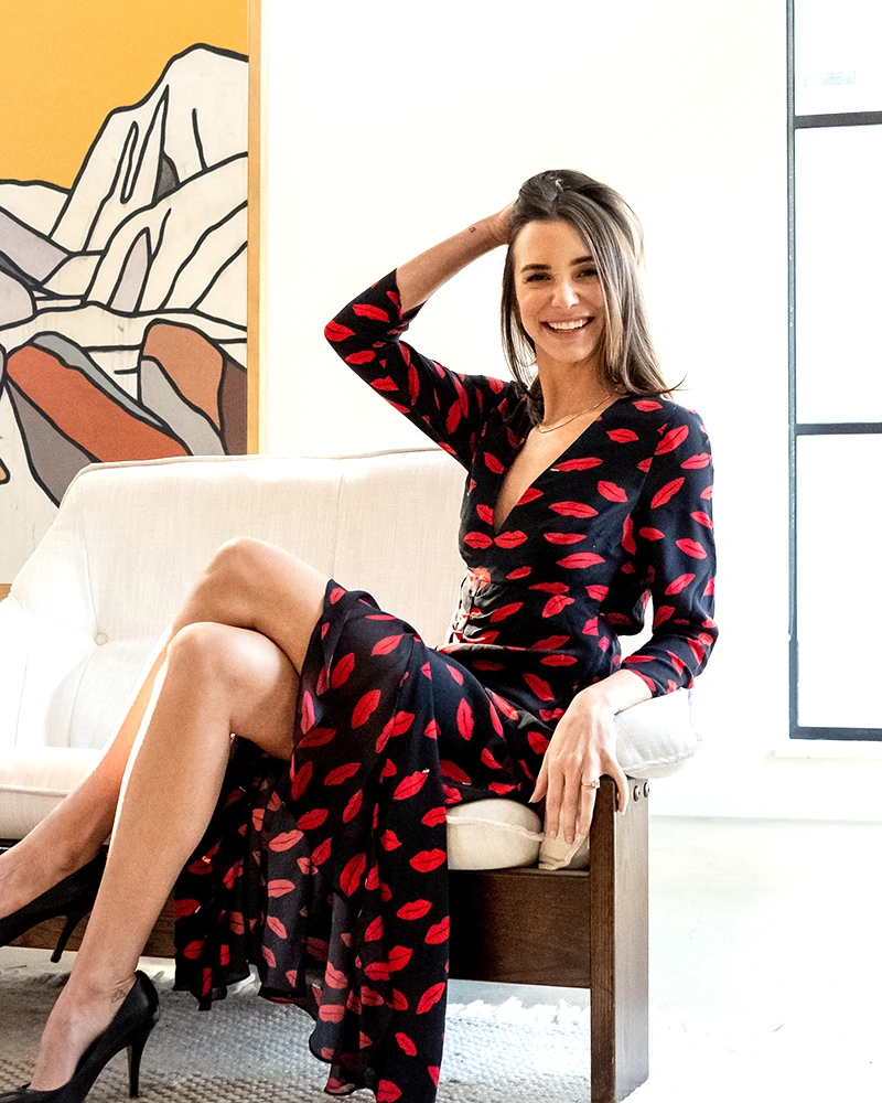 Photo of online dating coach Blaine Anderson, in a black dress with a red print, seated on a chair, with her hand on her hair, smiling